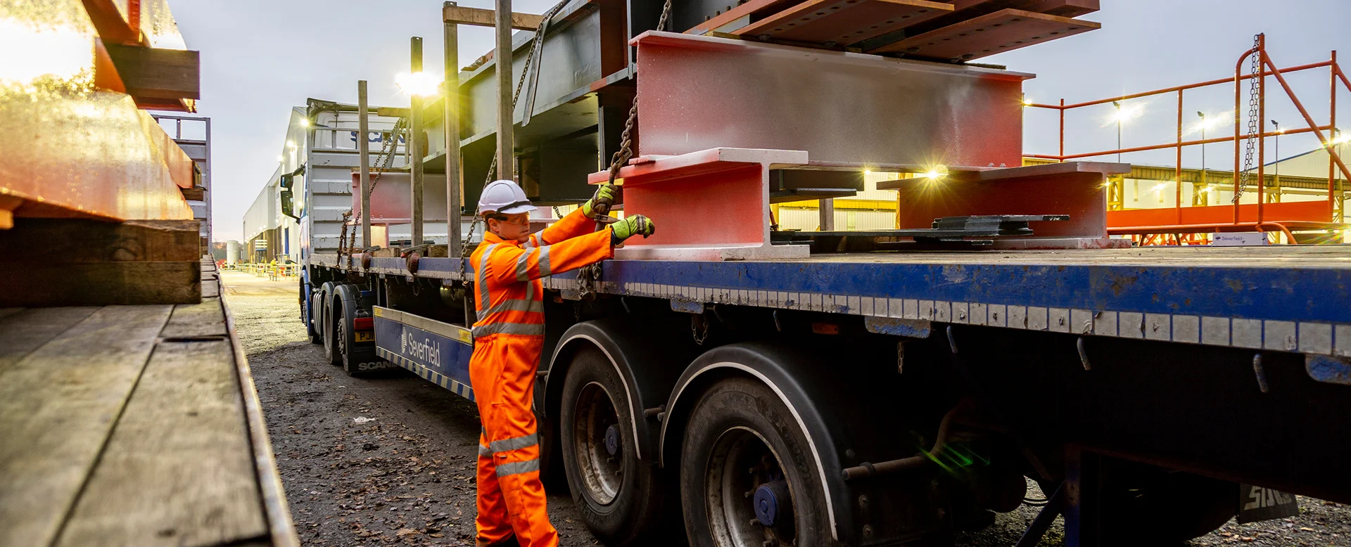 Driver Wearing Full PPE and Chaining Down a Load of Steel Structures onto a Semi Truck
