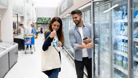 Man and woman grocery shopping down refrigeration aisle.