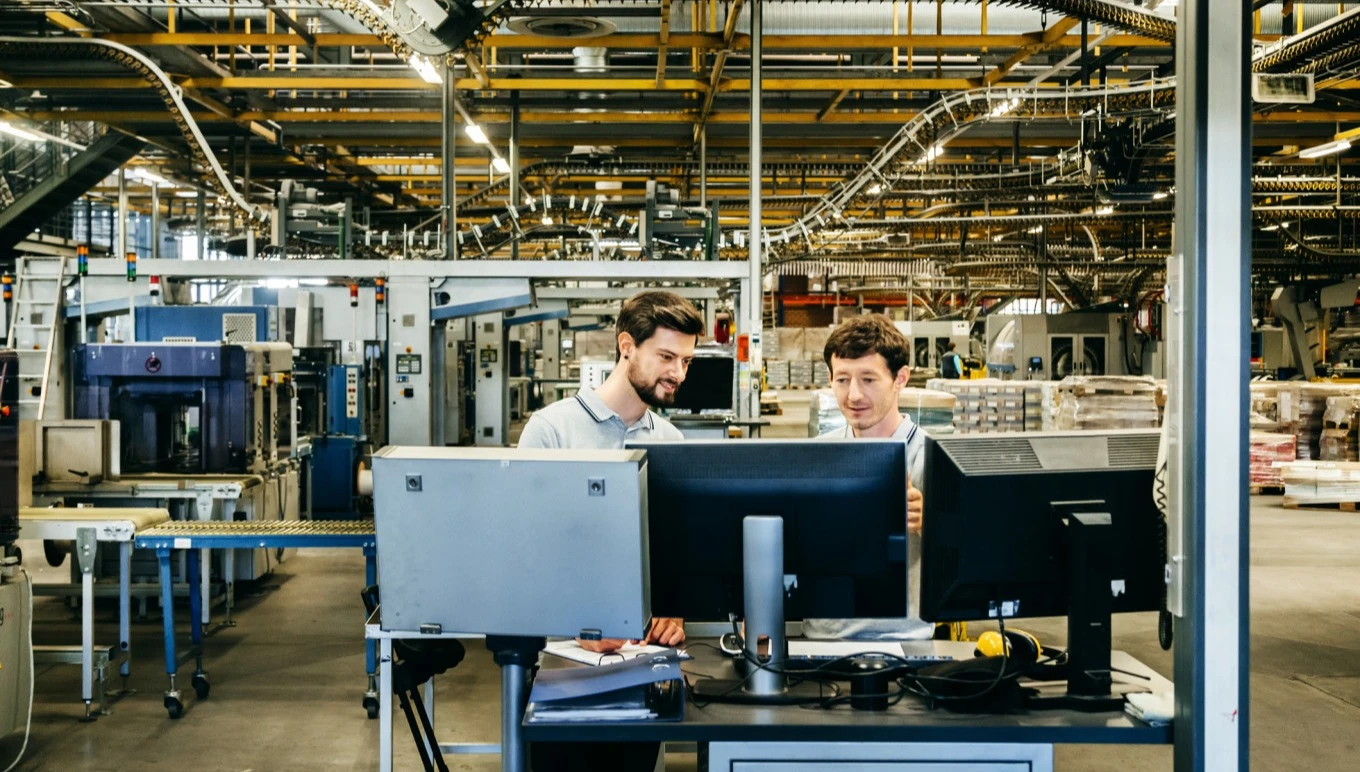 Engineers review data on monitors with manufacturing equipment in the background.