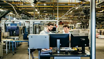 Engineers review data on monitors with manufacturing equipment in the background.