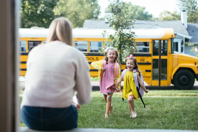 Two young girls running from the school bus across the front yard to their mother