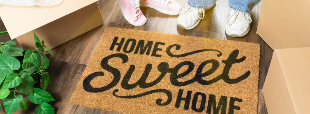 Home Sweet Home welcome mat surrounded by moving boxes and feet of two home owners