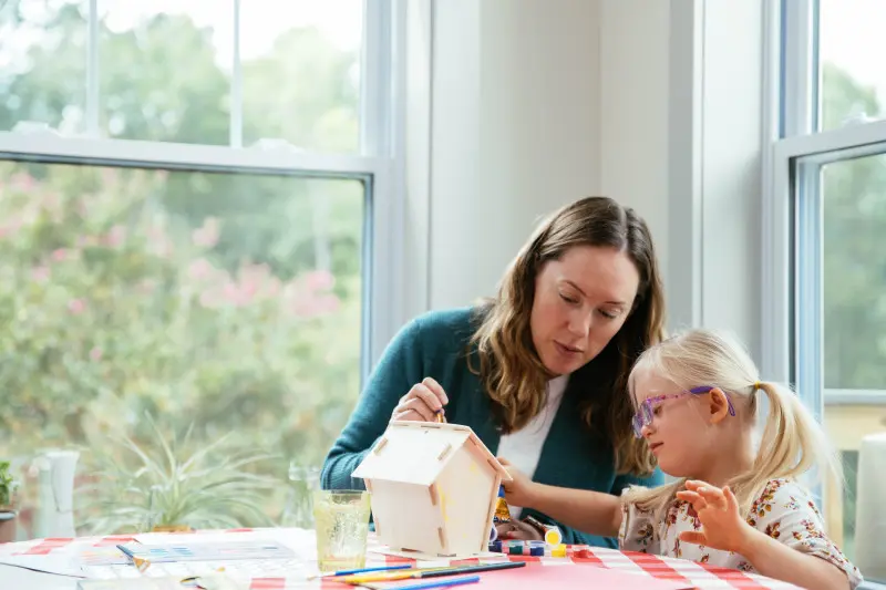 Mother painting birdhouse with her daughter at kitchen table