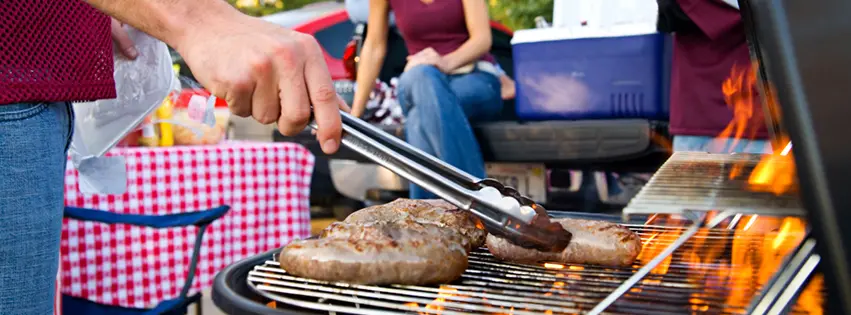 Person using tongs to grill sausages on grill at tailgate cookout before event