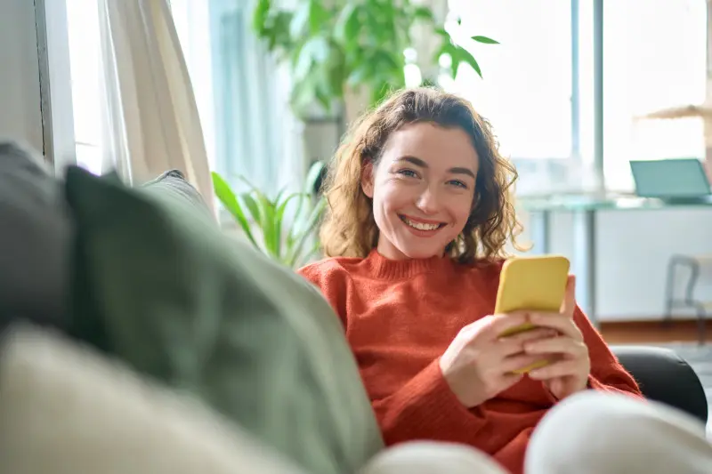 Woman wearing red shirt smiling and sitting on couch in her apartment that she rents