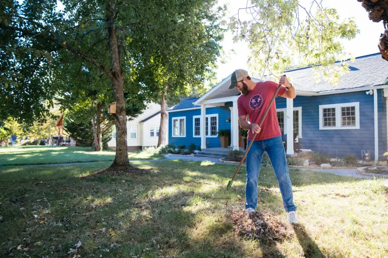 Tennessee homeowner in red tri-star shirt raking piles of leaves in front of the blue house that he owns