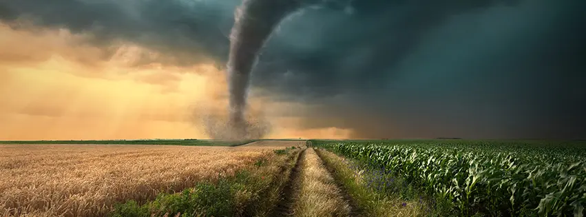 A large tornado with a swirling funnel cloud moving across a field. Debris is flying around the base of the tornado