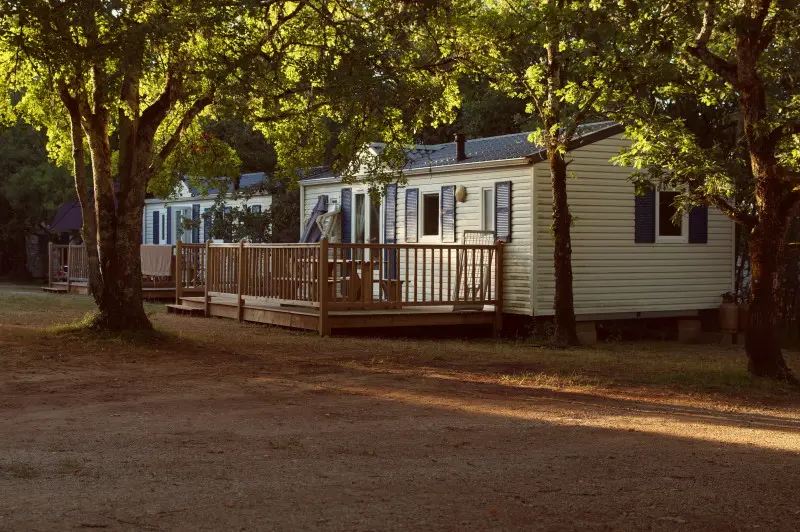 Mobile home with a wooden deck in a wooded area. There are trees with full green leaves surrounding the mobile home