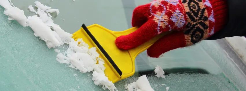 Hand wearing winter gloves scraping ice off a car windshield with a yellow ice scraper