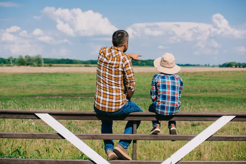 Father and son rest on fence while father points out something interesting in field