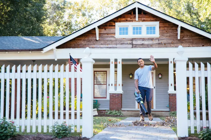 Homeowner waving to friend while blowing leaves off the front lawn that is surrounded by a white fence