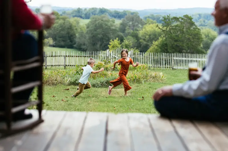 Two kids running and playing in the front yard while their grandparents watch from the front porch