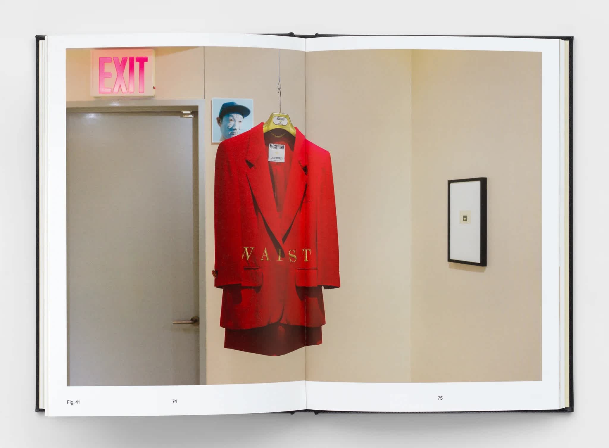 Open book with centerfold exhibition image. A red coat hangs in the center. A frame hangs on the wall to the right of the coat. A door and exit sign above it, are to the left of the coat.