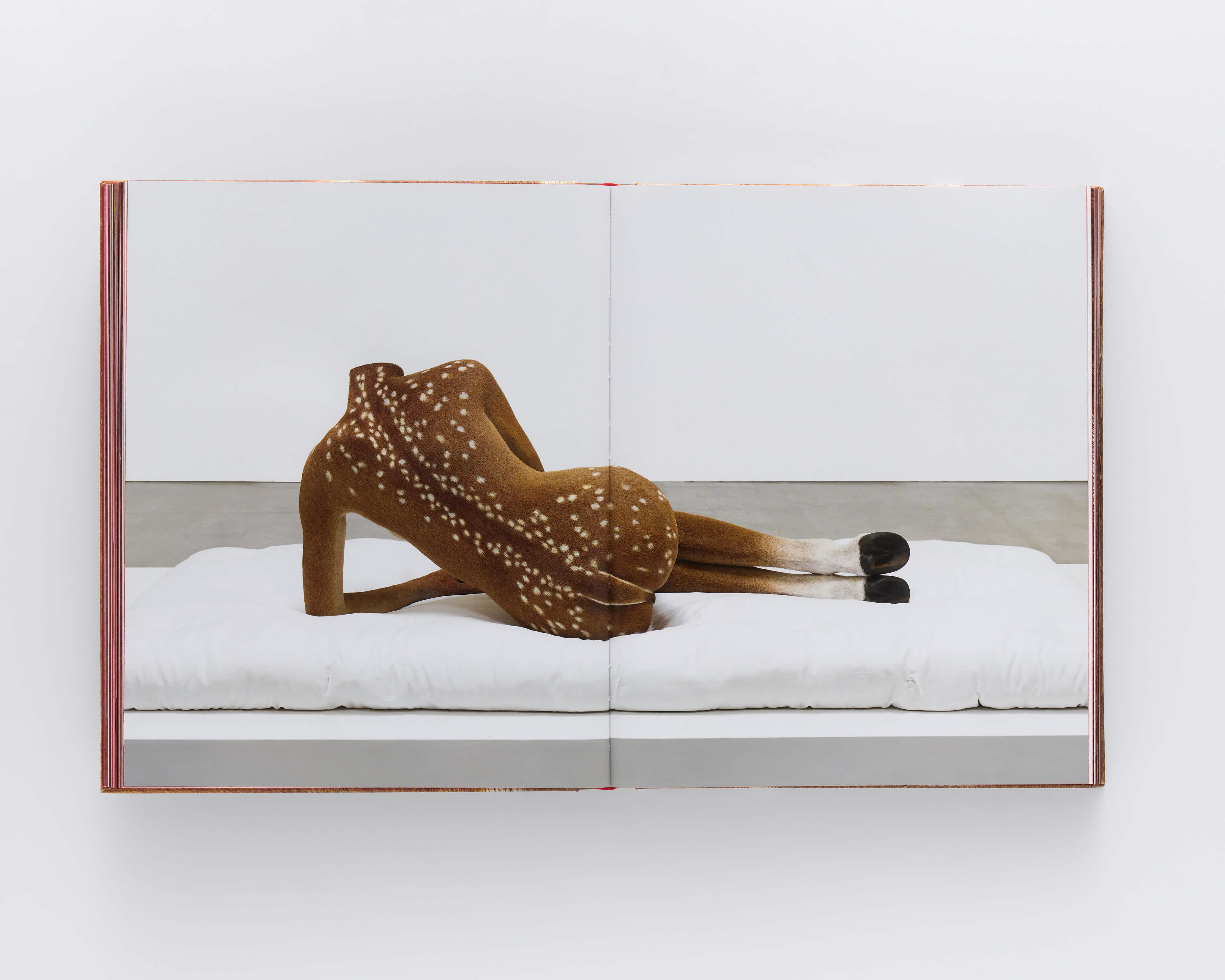 Open book with full-bleed centerfold image of a sculpture of a headless woman. It lays on top of a white cushion, facing away from the viewer. Instead of skin the sculpture is covered in a speckled-fawn's coat of fur. Instead of feet, the sculpture has hooves.
