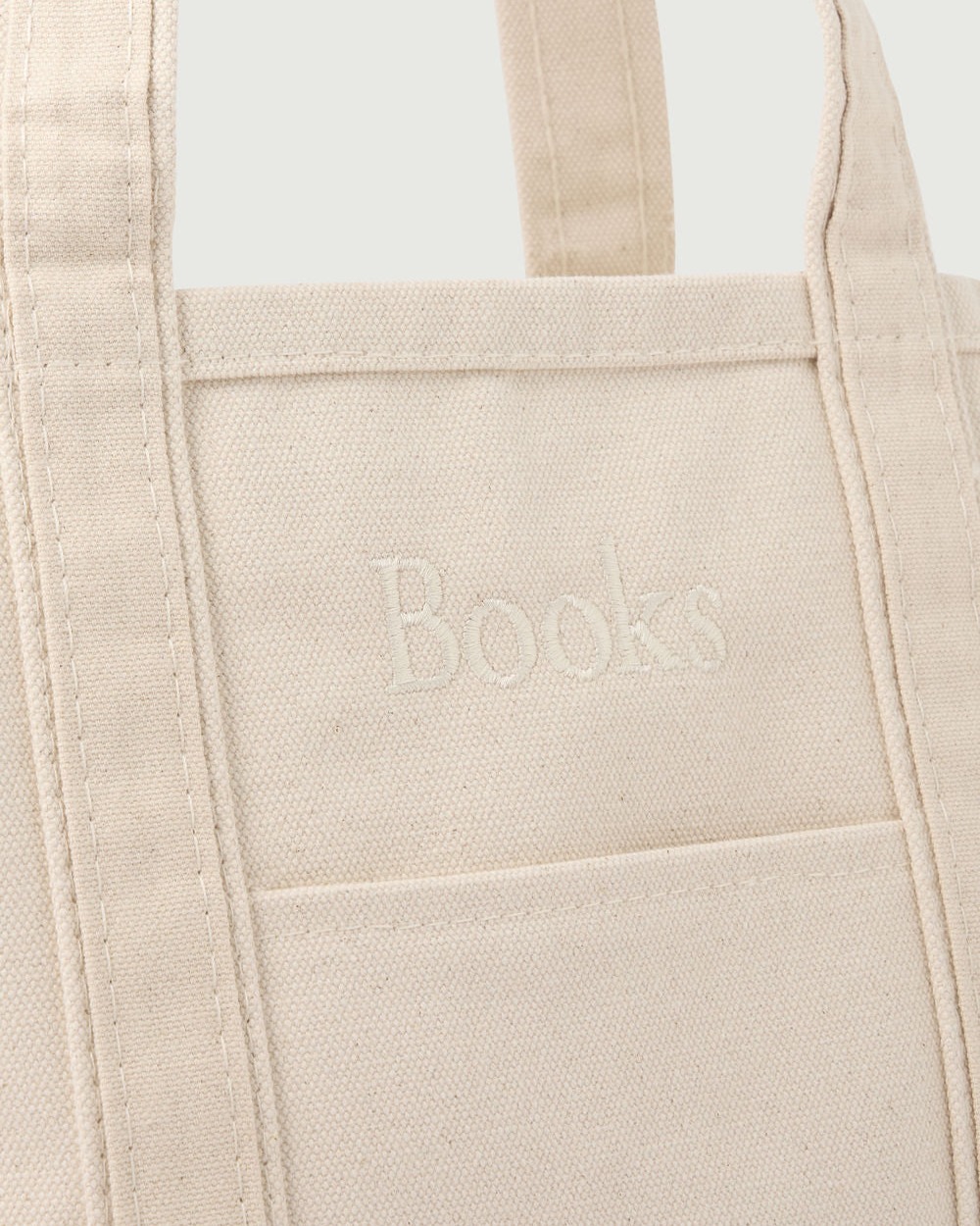 A close-up of a beige canvas tote bag with the word Books embroidered in white above a front pocket.