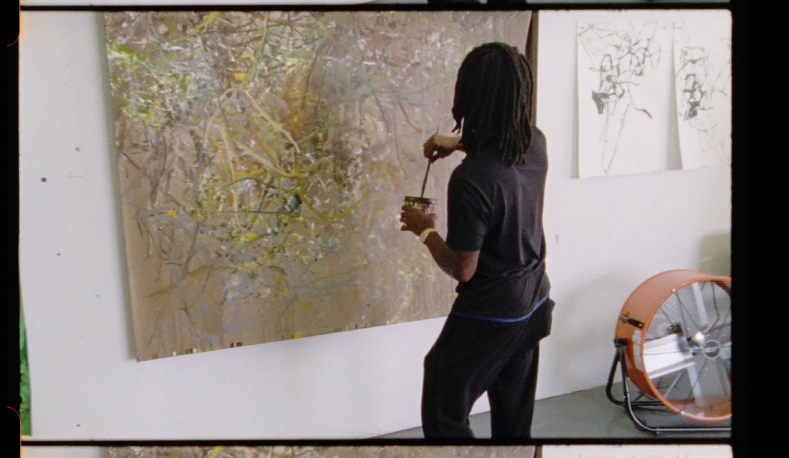 An artist faces a canvas on a wall. His back is to us. In his left hand he holds a can of paint. His right hand dips a brush into the paint.