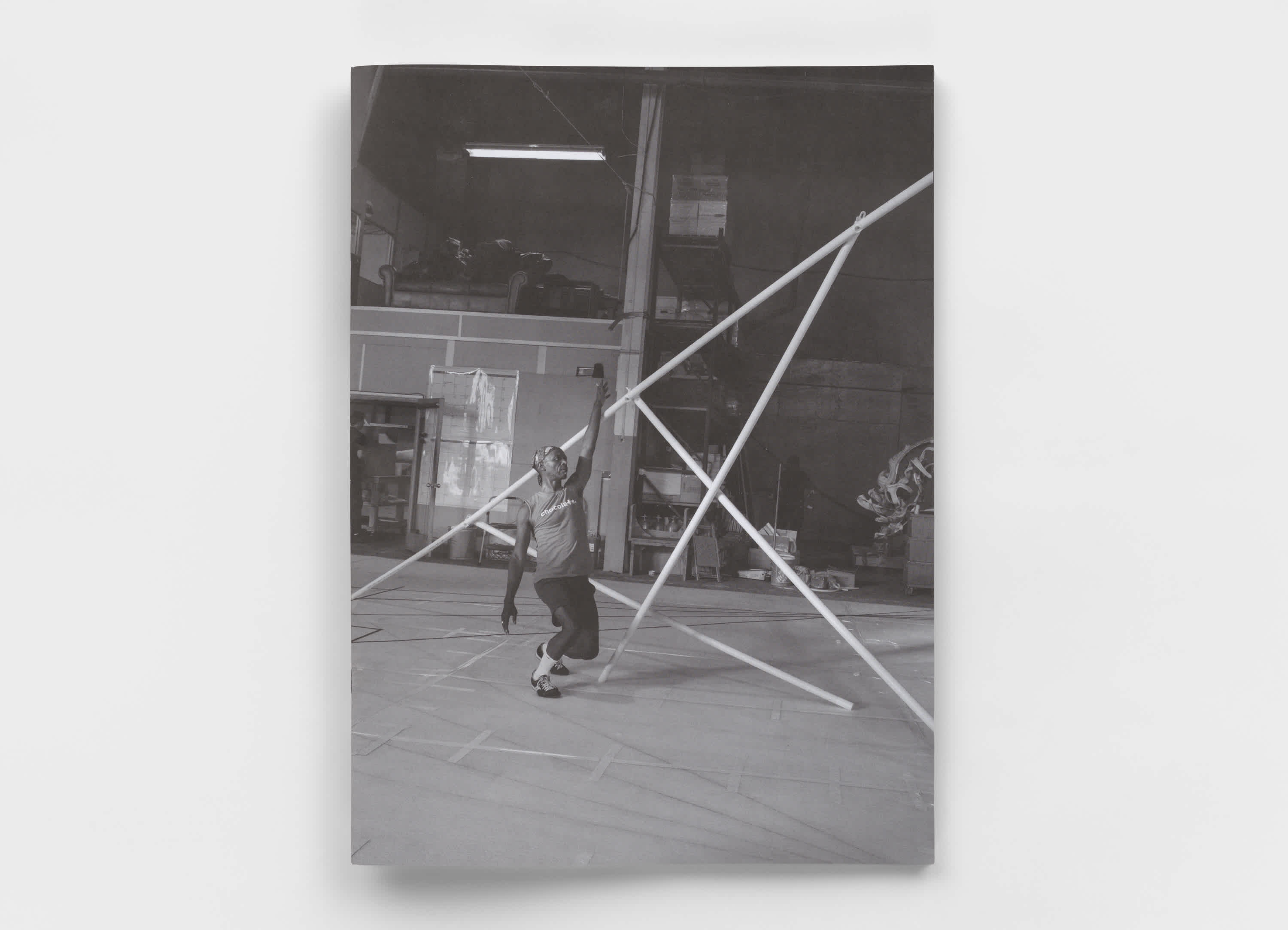 Black and white book cover is on top of a gray background. The cover features a dancer on his way toward taking a knee. His fist is raised and he is positioned in front of four criss-crossing white PVC poles. A large gymnastics mat covers the floor of the warehouse room he is in.