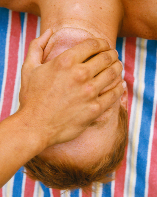 A person with short, light brown hair lies on a colorful striped towel while another hand covers their mouth and chin. The scene is brightly lit, suggesting an outdoor or sunny setting.