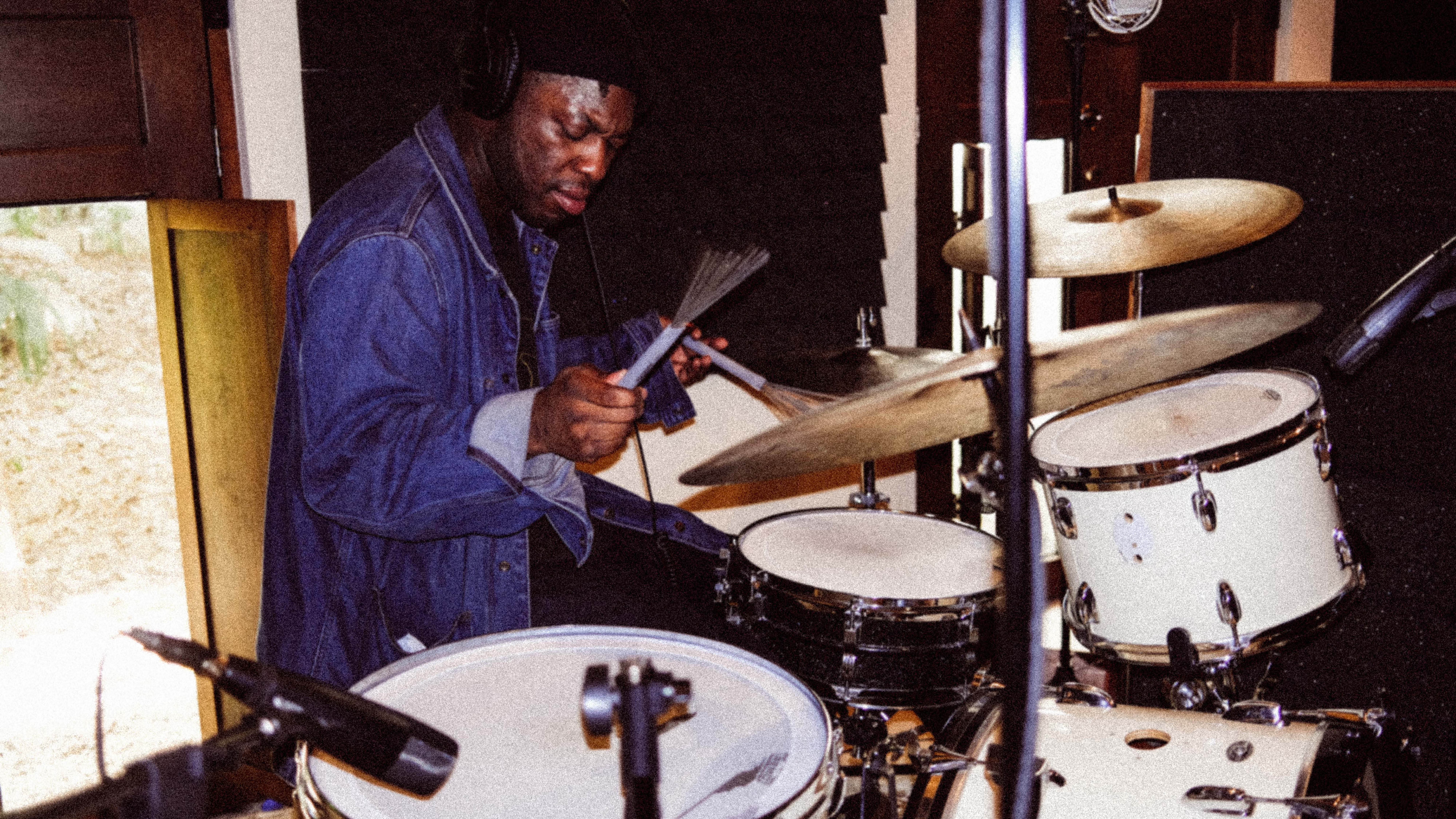 A man wearing headphones and a denim jacket plays a white drum set in a music studio, focusing intently on the drums while holding drum brushes. Microphones and soundproofing panels are visible around him.