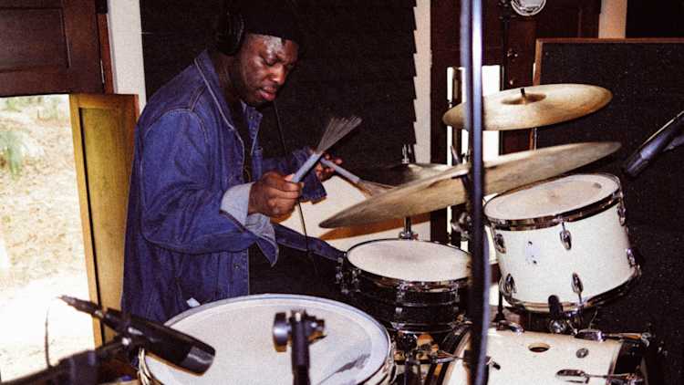 A man wearing headphones and a denim jacket plays a white drum set in a music studio, focusing intently on the drums while holding drum brushes. Microphones and soundproofing panels are visible around him.