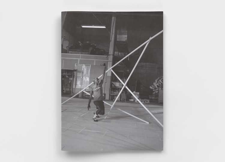 A black-and-white photo shows a person balancing on one foot while holding a tall, angular structure made of thin rods in what appears to be an industrial workshop or studio space.