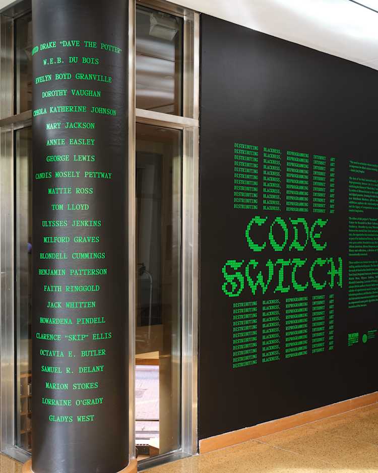 A museum wall displays the words CODE SWITCH in large green text, surrounded by a grid of names, with more names listed vertically on a glass panel beside the wall. The design mimics green computer code.