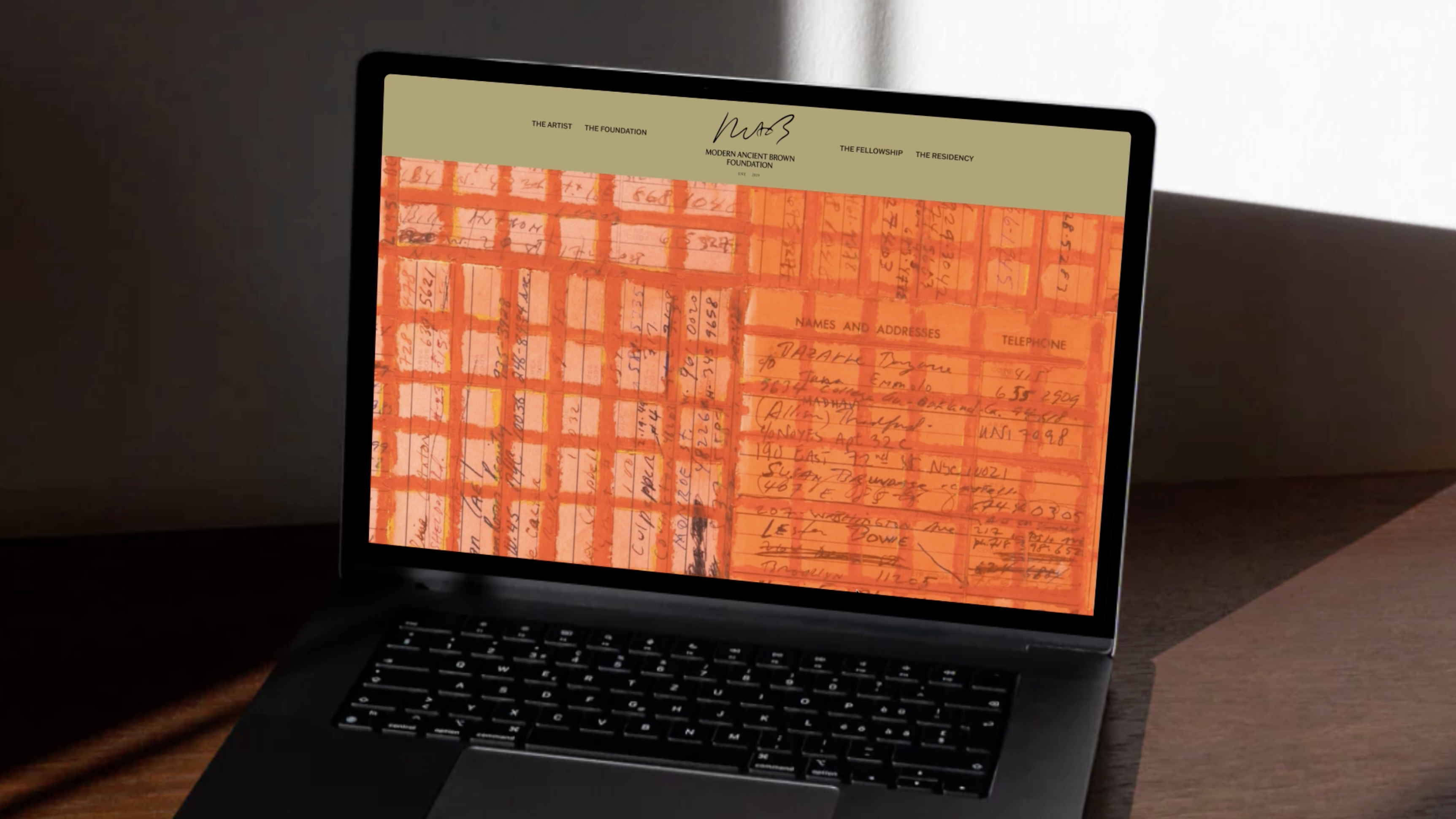 A laptop on a dark table displays a website with an orange background featuring handwritten notes, names, and phone numbers. Sunlight casts shadows on the table and wall behind the laptop.