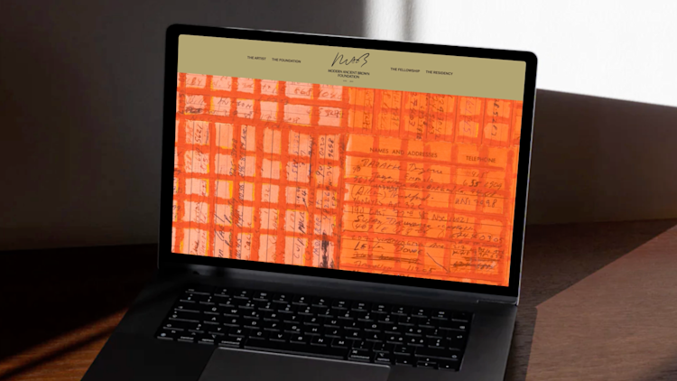 A laptop on a dark table displays a website with an orange background featuring handwritten notes, names, and phone numbers. Sunlight casts shadows on the table and wall behind the laptop.