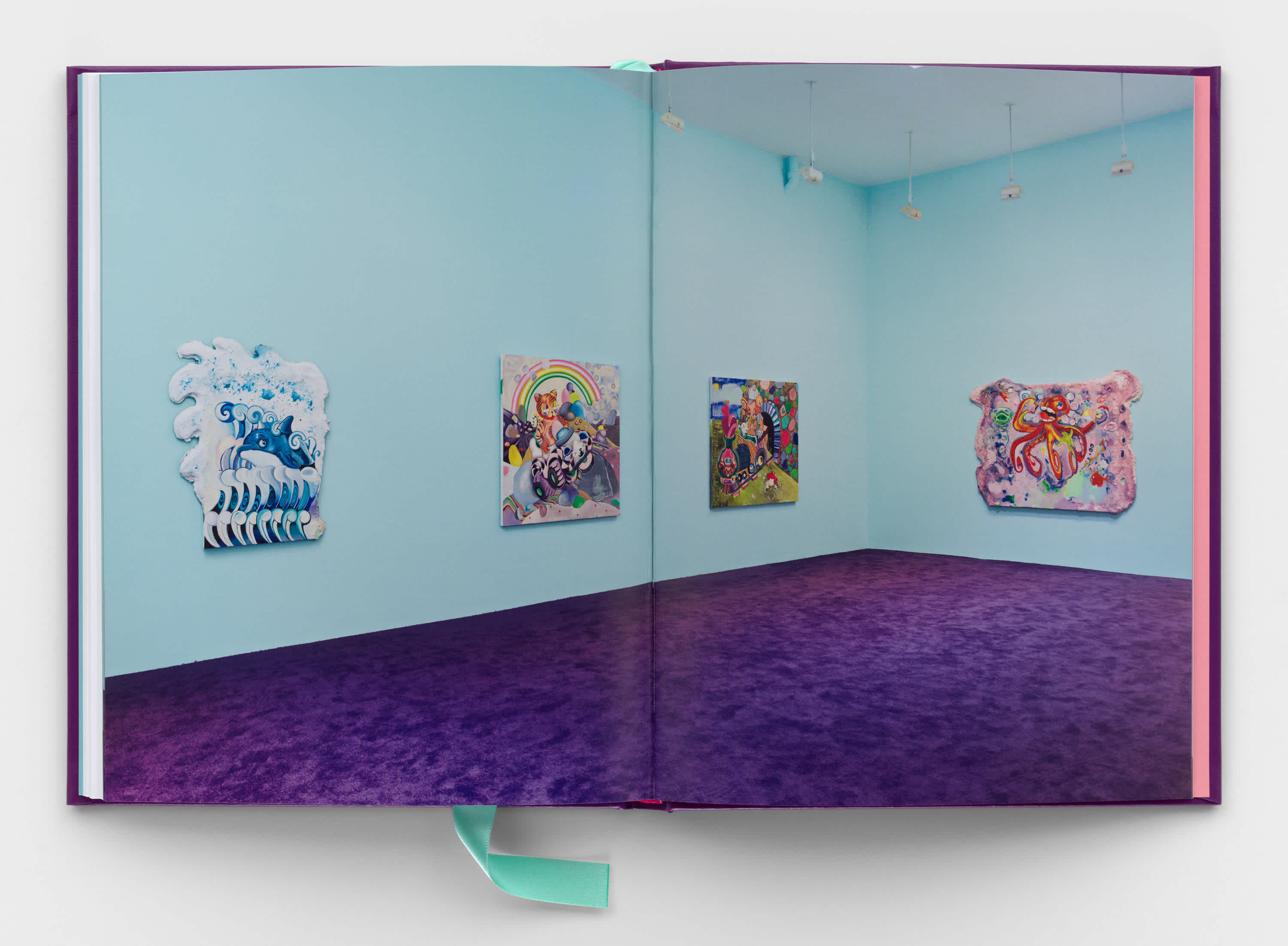 A book centerfold that shows a full bleed image of an exhibition. It's a corner of a room in which four colorful paintings hang on a light blue wall. The carpet on the floor is deep purple.