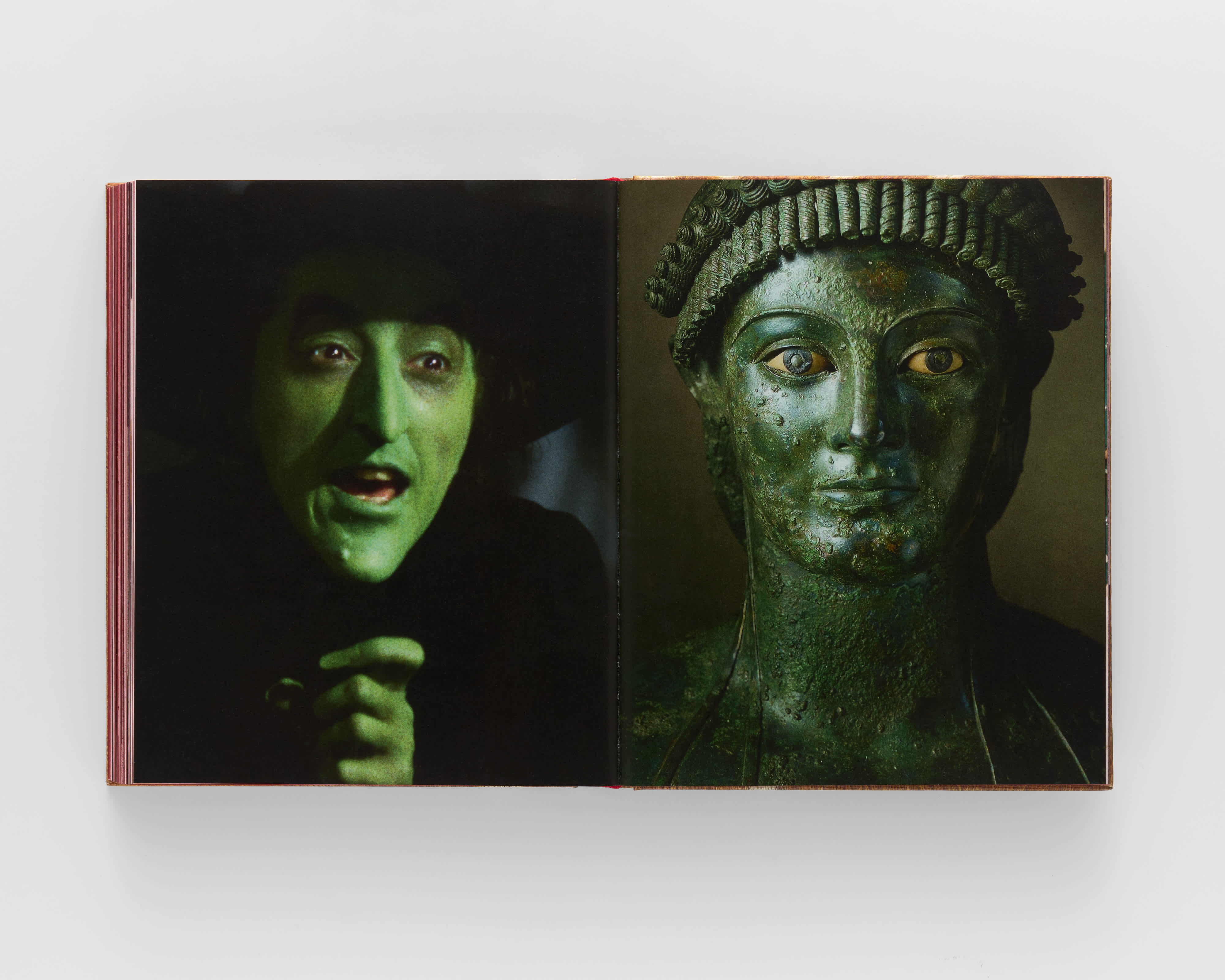 An open book on a grey background. The left page features a photograph of The Wicked Witch of The West from the original Wizard of Oz film. The right page juxtaposes a metal bust of a woman, turned green with age, from the Hellenistic period. 