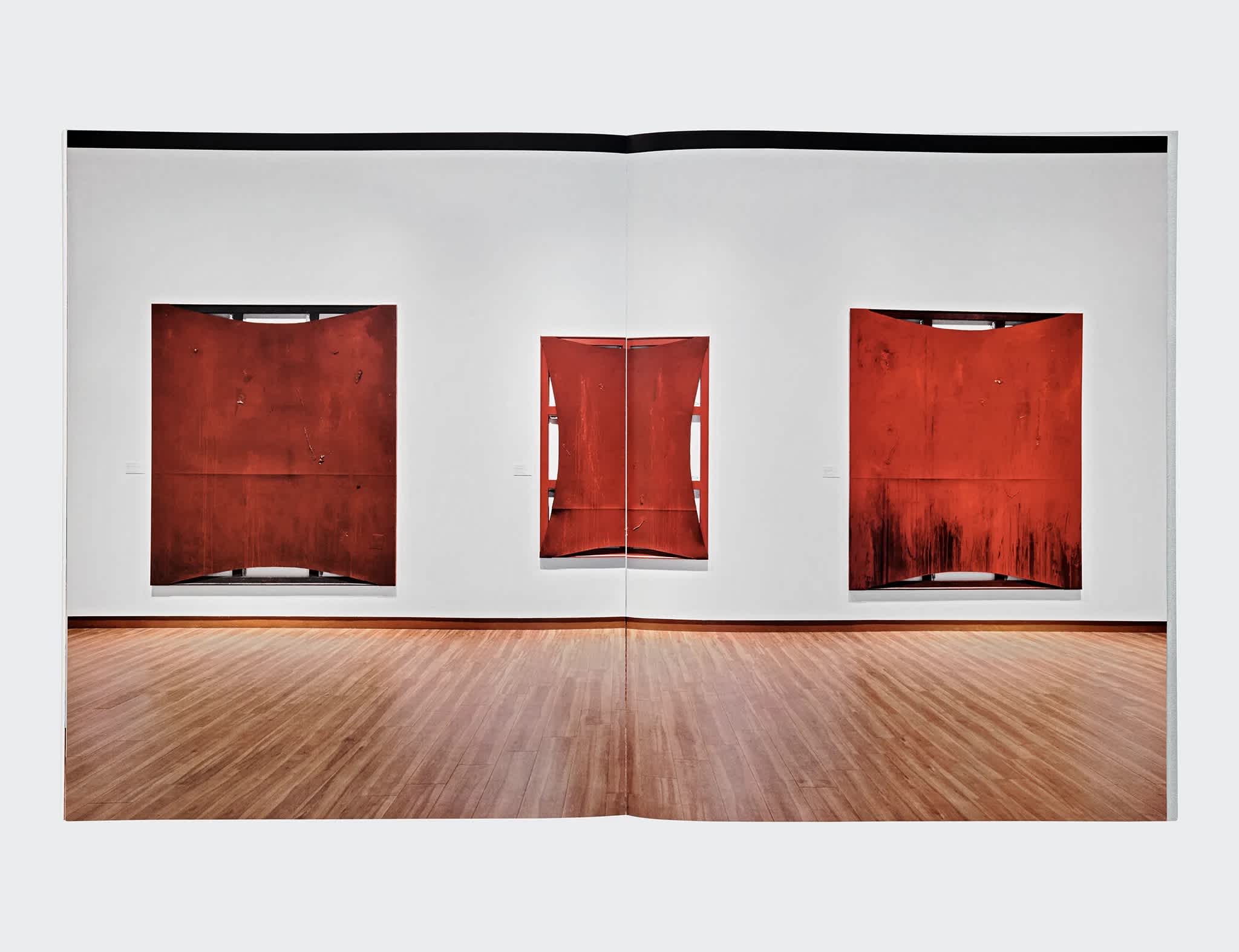 Book interior that features a centerfold of three red paintings hung on a white wall. The floor in front of the wall is made of wood planks. 