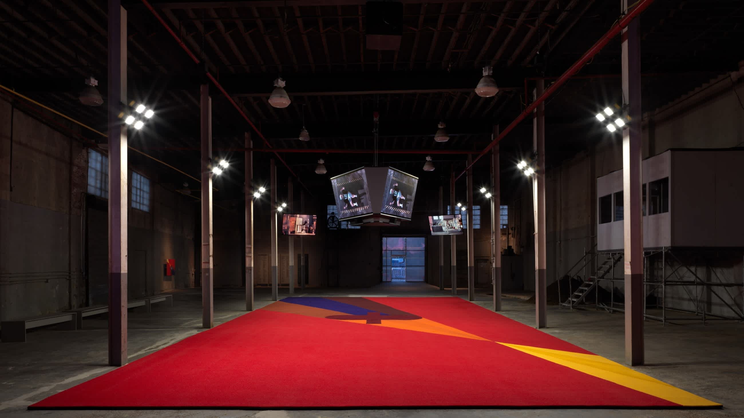 Still photo from the artist Matthew Barney's installation, 'Secondary'. A red, orange and blue carpet sits between rows of pillars within a warehouse. A sports jumbotron is suspended above the carpet. Football field lights are attached to the tops of the pillars. Barney has created a carpeted football field within a warehouse.