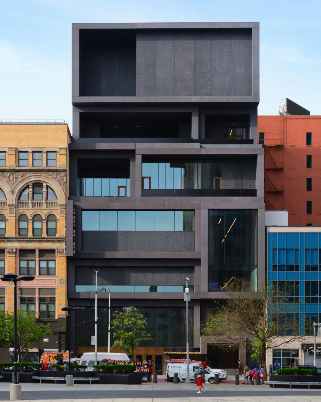 A modern, boxy building with dark gray stacked levels and large glass windows, standing between older, lighter brick buildings. People and vehicles are visible in front on the street.