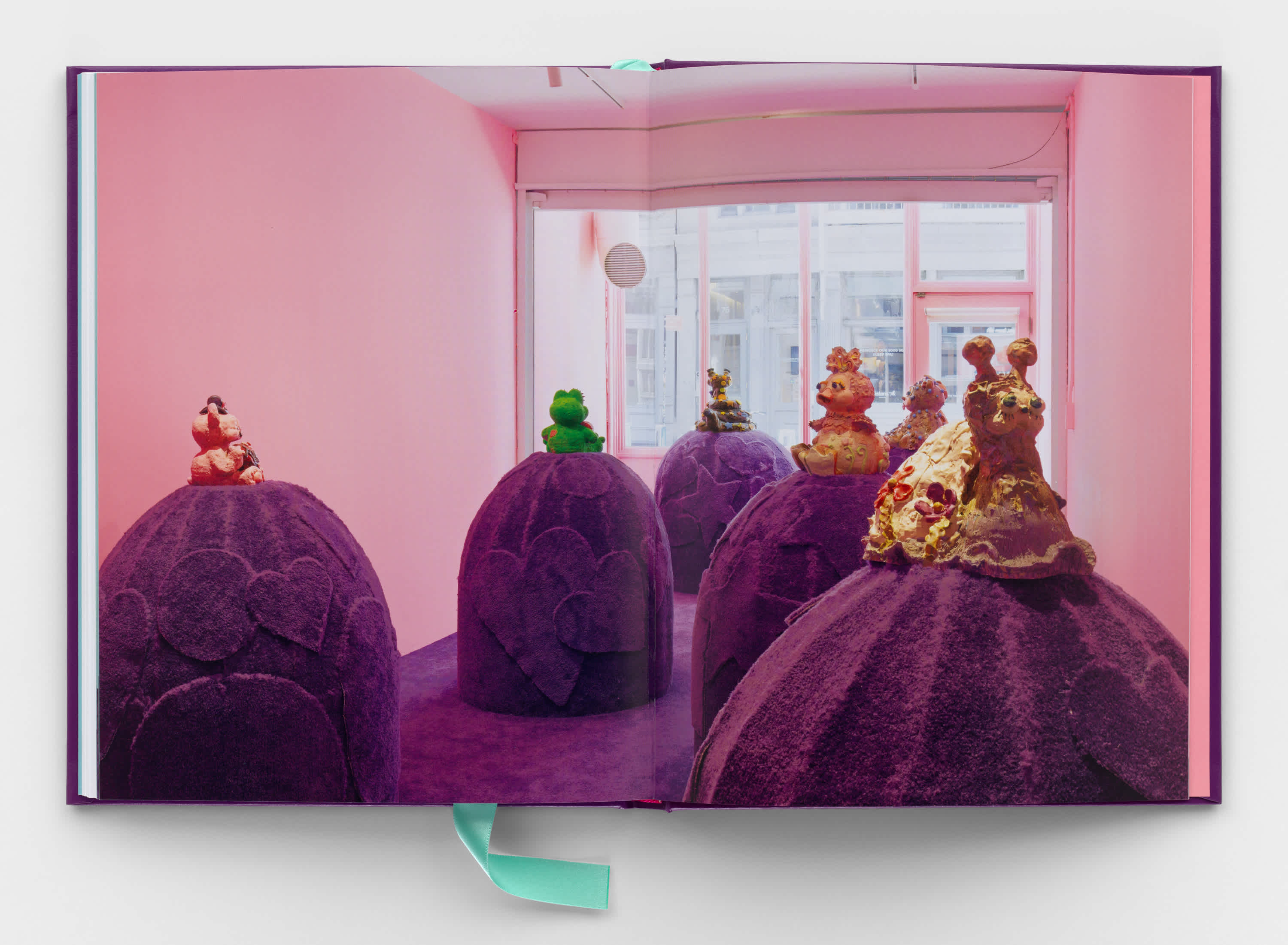 A book centerfold that shows a full bleed image of an exhibition. Several large purple carpet mounds rise up out of the floor. A colorful cartoon creature sits atop each mound. A wall of windows in the background looks out onto the street. The walls are pink.