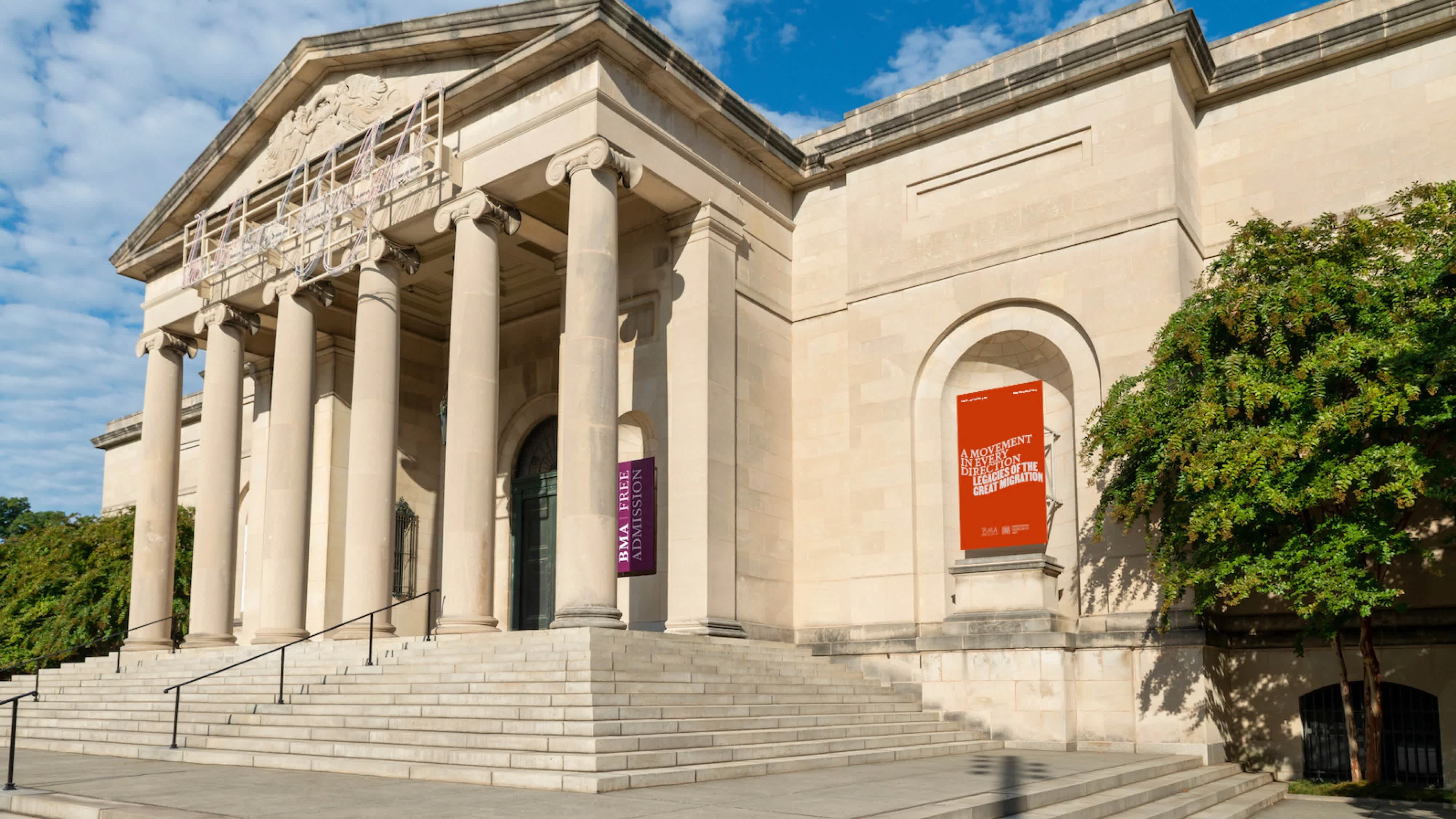 Greek style stone building with stairs leading up to an entrance just behind a row of ionic columns. A red banner sits on the exterior of the building to the right of the stairs.