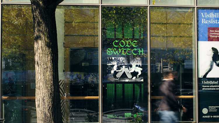 A glass window displays a green digital-style sign reading CODE SWITCH above black-and-white images and green text columns, with a blurred person walking by and a tree partially shading the scene.