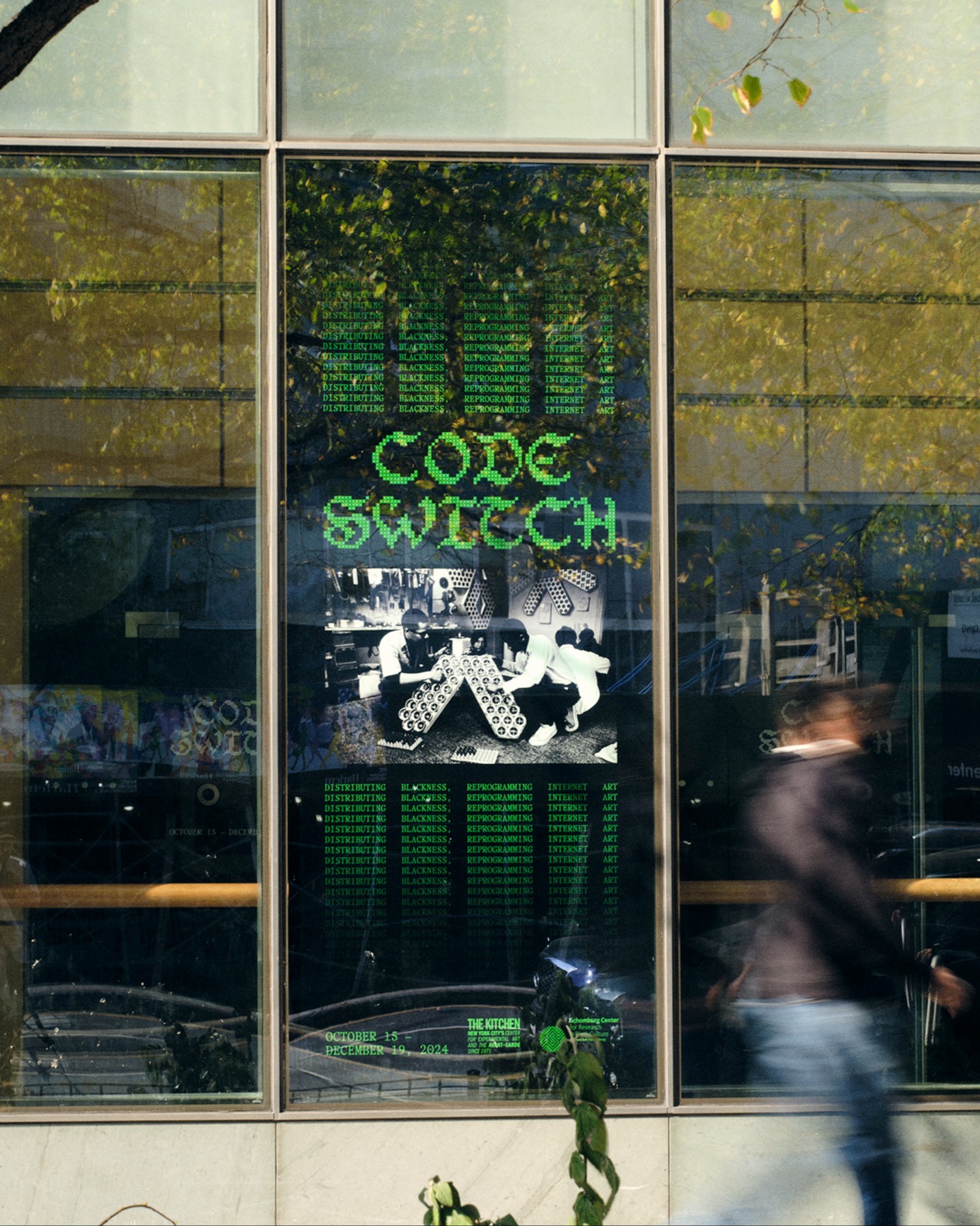 A blurred person walks past a large window displaying digital green code, the words CODE SWITCH, and a black-and-white image of people playing chess. Reflections of trees and buildings are visible on the glass.