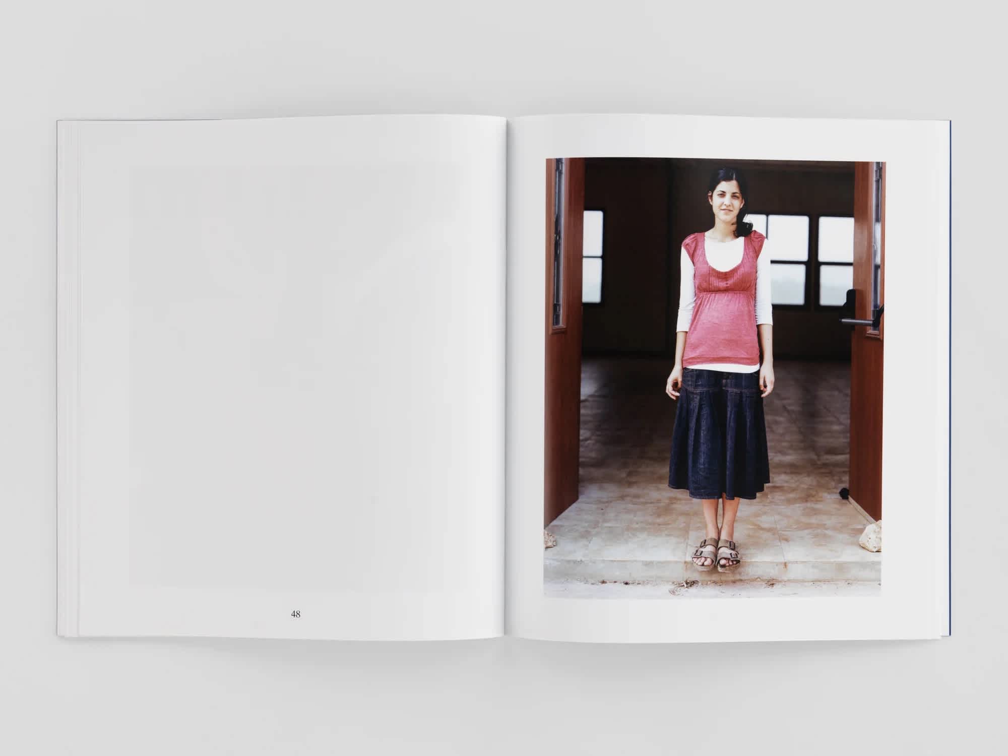 An open book. The left page is blank, the right page has a photograph of a woman wearing a pink shirt and black skirt, standing in a doorway.