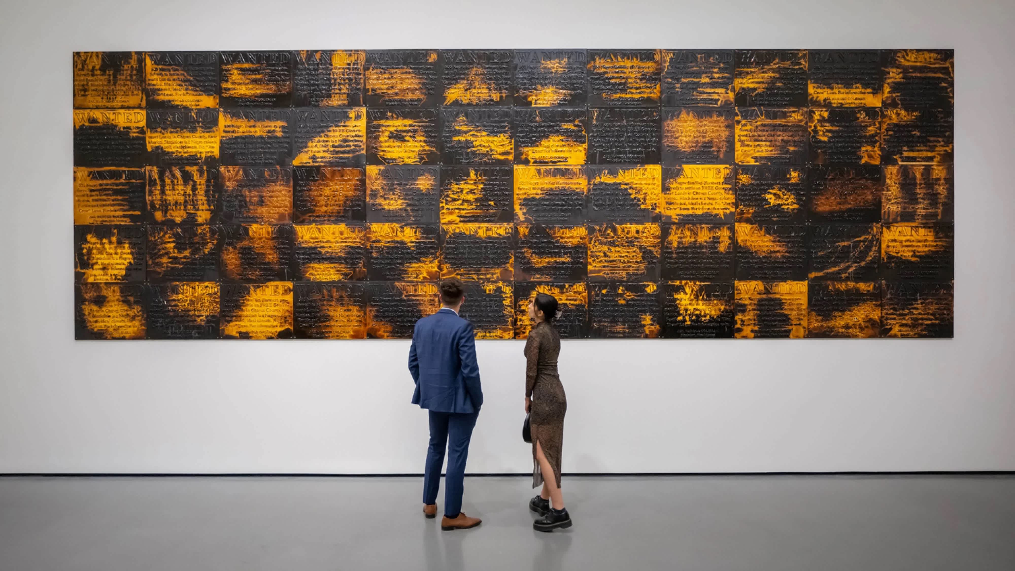 Two people stand in front of a large black and orange artwork. Their backs are to the camera and their heads are titled up towards the work.