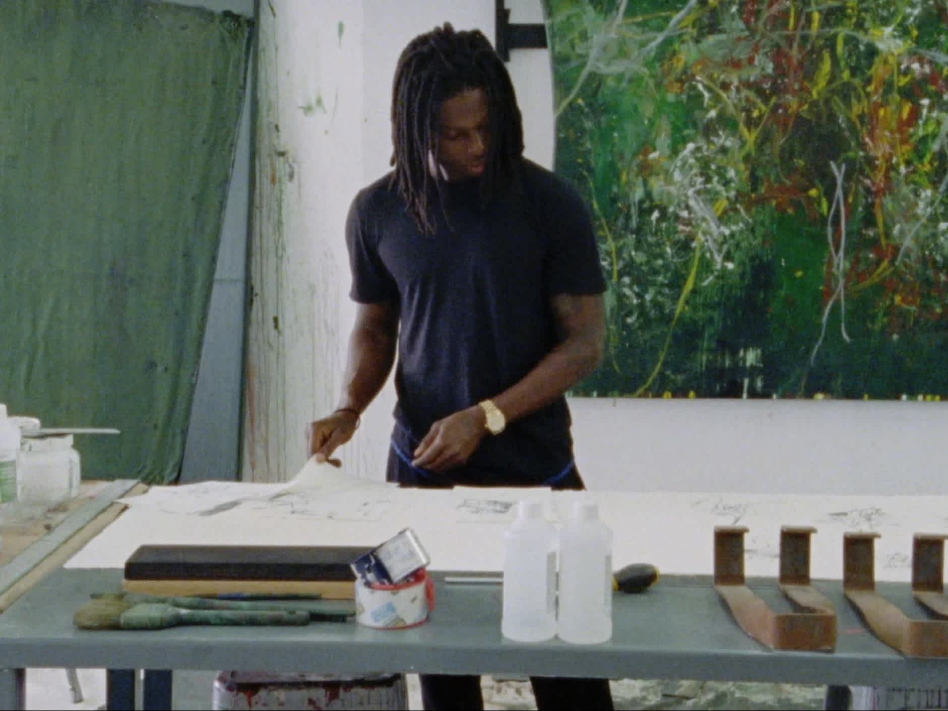 An artist stands at a table making work. Large abstract paintings are behind him on the wall.