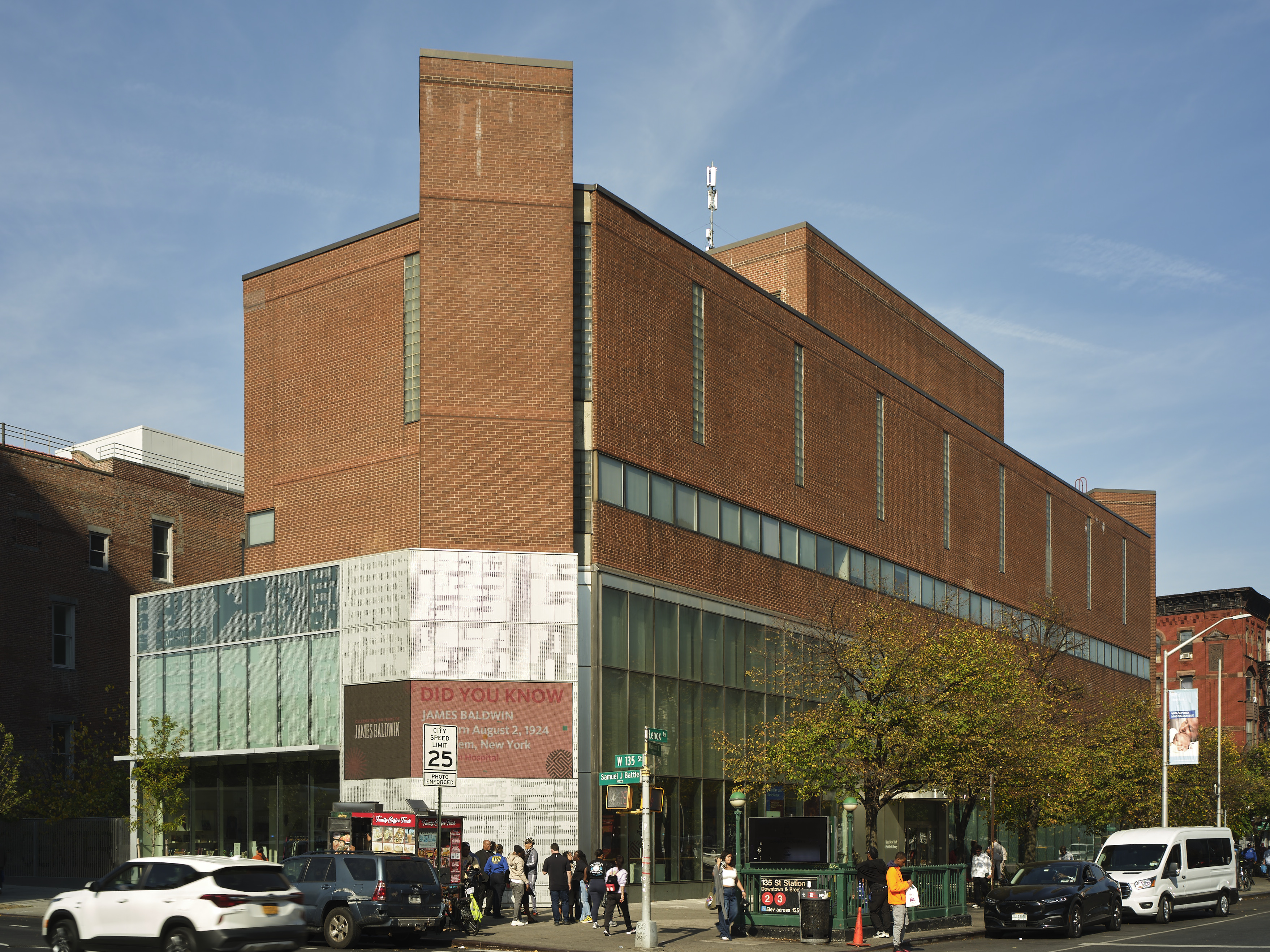 A modern red brick building with large windows on the ground floor, located on a street corner. Several people are gathered near the entrance, and cars are parked along the street under a clear sky.