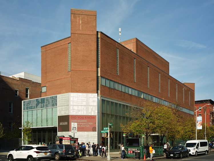 A modern red brick building with large windows on the ground floor, located on a street corner. Several people are gathered near the entrance, and cars are parked along the street under a clear sky.