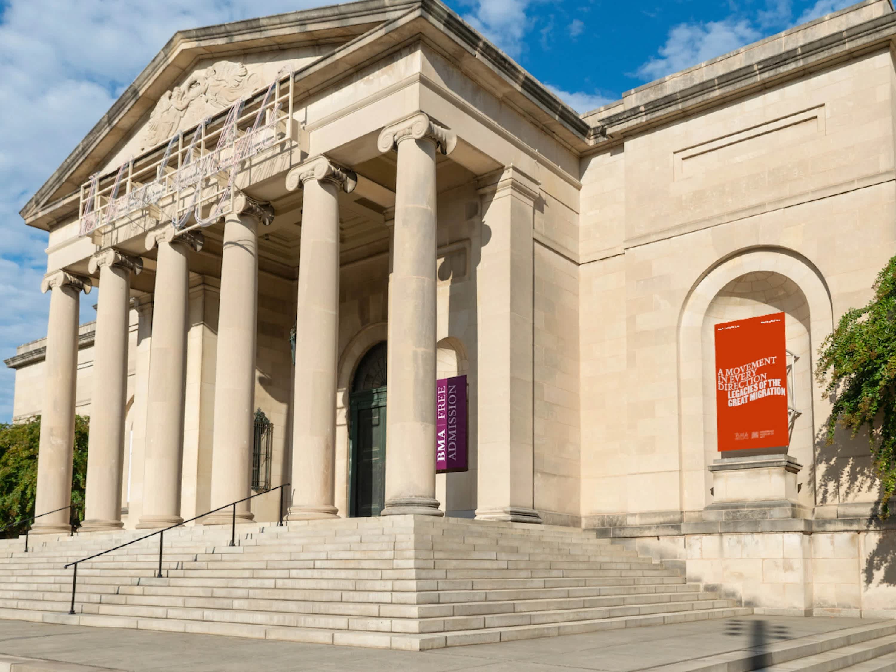 Greek style stone building with stairs leading up to an entrance just behind a row of ionic columns. A red banner sits on the exterior of the building to the right of the stairs.
