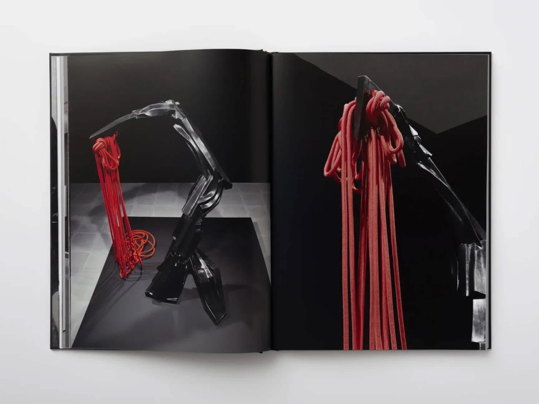 Two black metal sculptures holding long red rope in black rooms. The images of each sculpture are on opposite pages of a book.