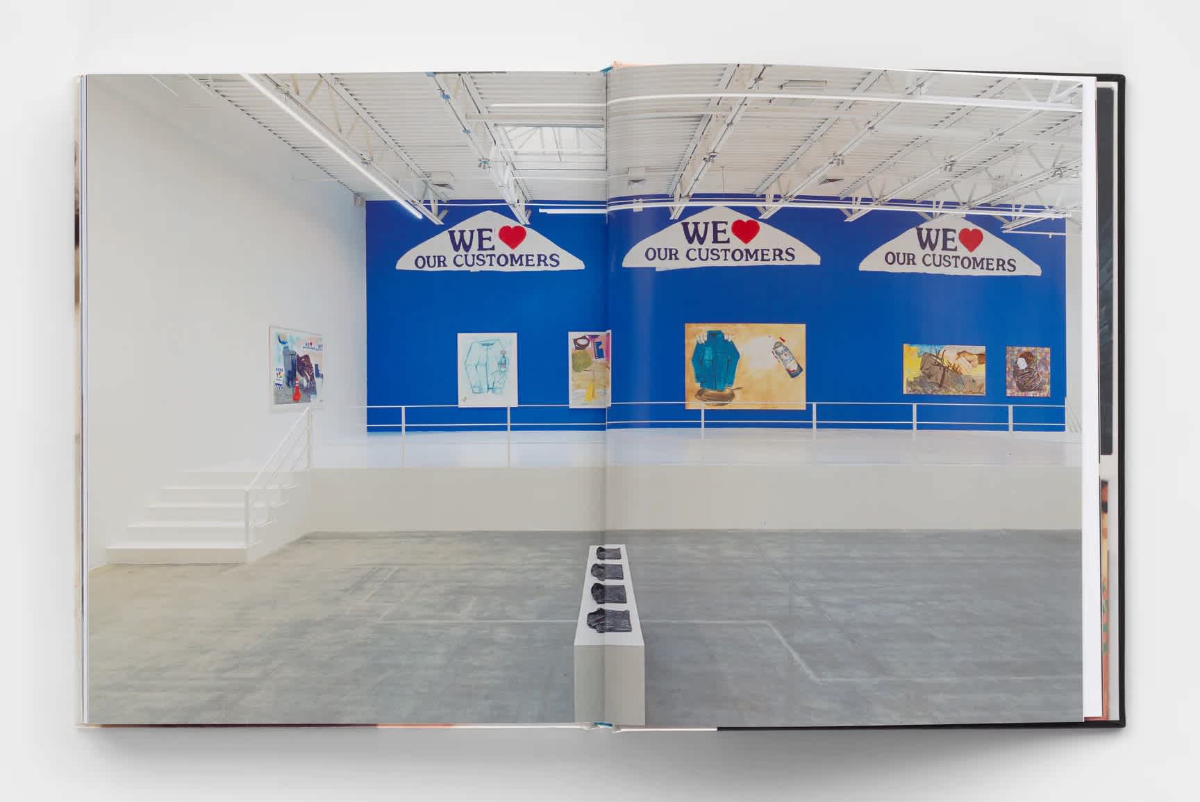 Book interior which features an installation shot from Pat Phillips' show at Jeffrey Deitch titled, "Consumer Reports". The room is mostly empty except for a white pedestal in the center which displays four folded shirts. A set of stairs on the left of the room leads up to a railed platform. The wall behind the platform is painted bright blue and has five paintings hung beneath three large-scale murals of dry-cleaning hangers. 