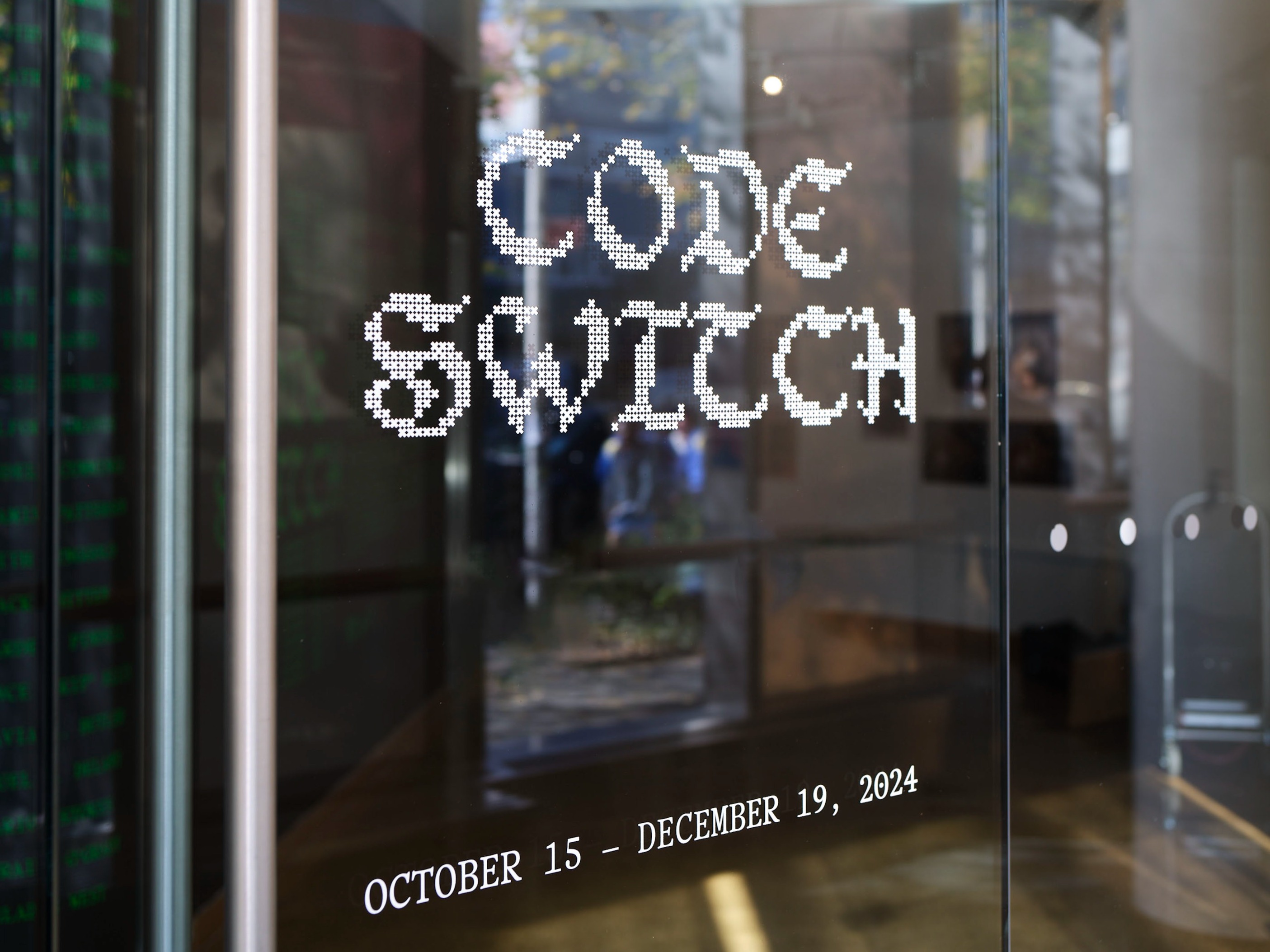 Glass door with the words “CODE SWITCH” in pixelated font and event dates “OCTOBER 15 – DECEMBER 19, 2024” in white text, reflecting the interior of an art gallery or exhibition space.