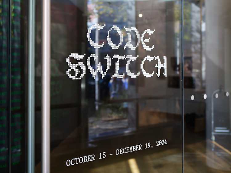 Glass door with the words “CODE SWITCH” in pixelated font and event dates “OCTOBER 15 – DECEMBER 19, 2024” in white text, reflecting the interior of an art gallery or exhibition space.