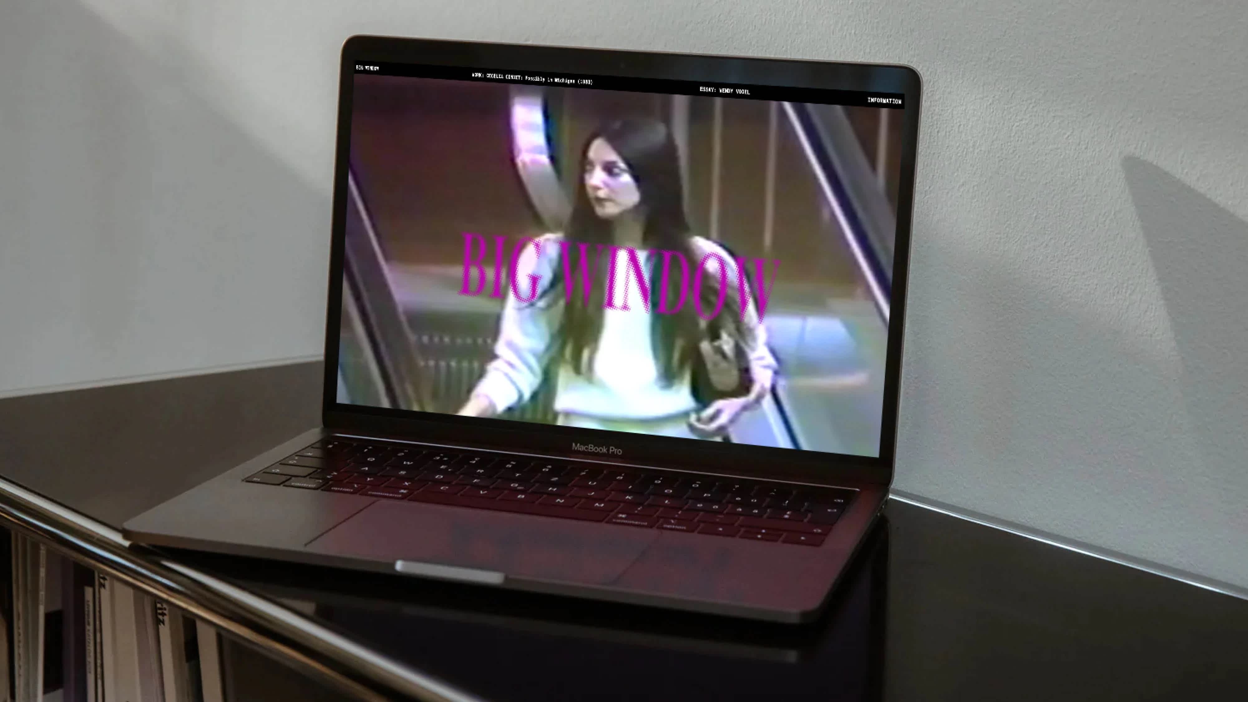 A laptop rests on top of a black bookshelf. The screen features a woman going down an escalator. The title "Big Window" stretches across the screen in magenta, serif letters.