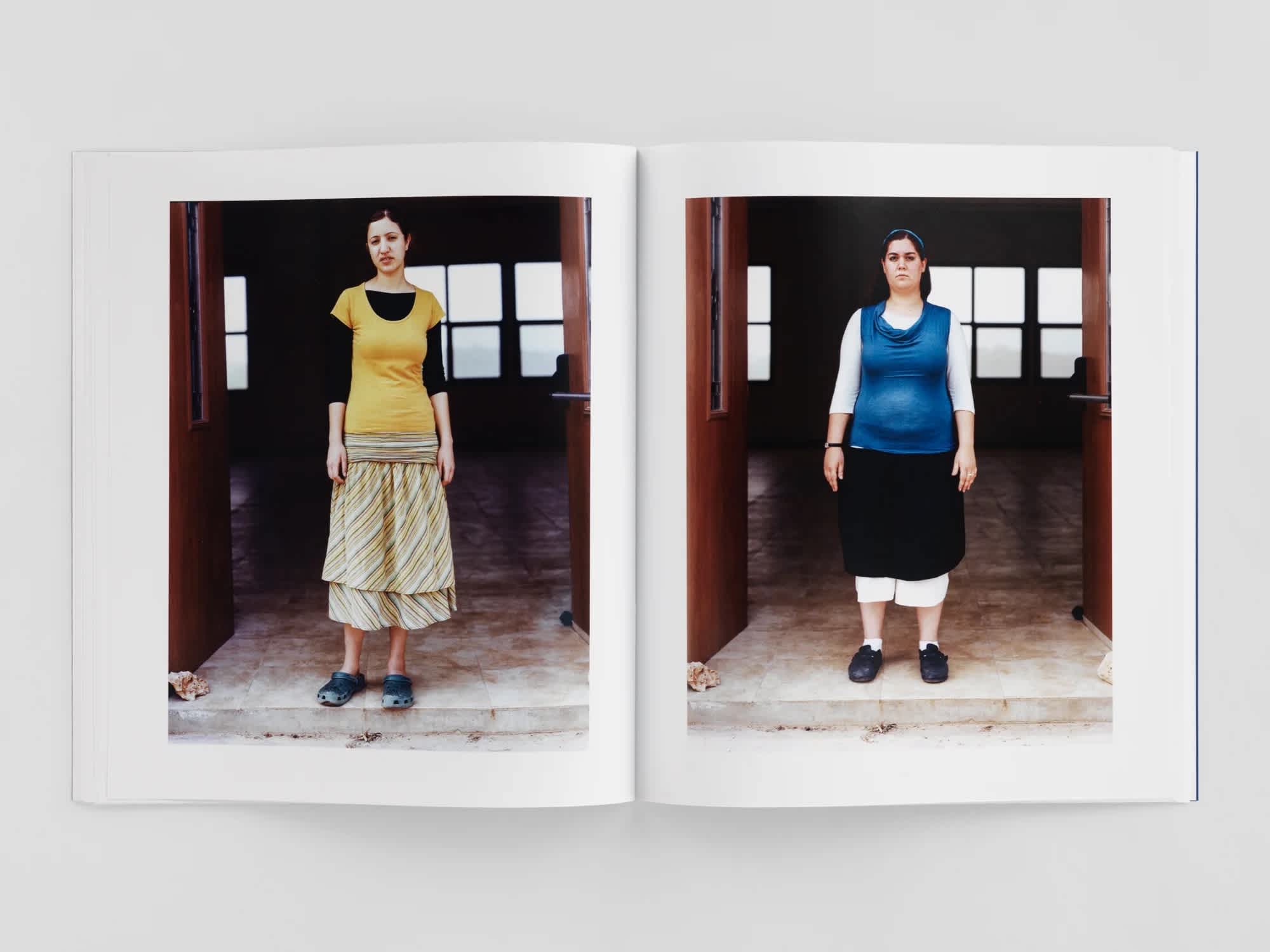 An open book. Each page has a photograph of a woman standing in a large doorway. The woman on the left page wears mostly yellow. The woman on the right wears a blue shirt and black skirt.