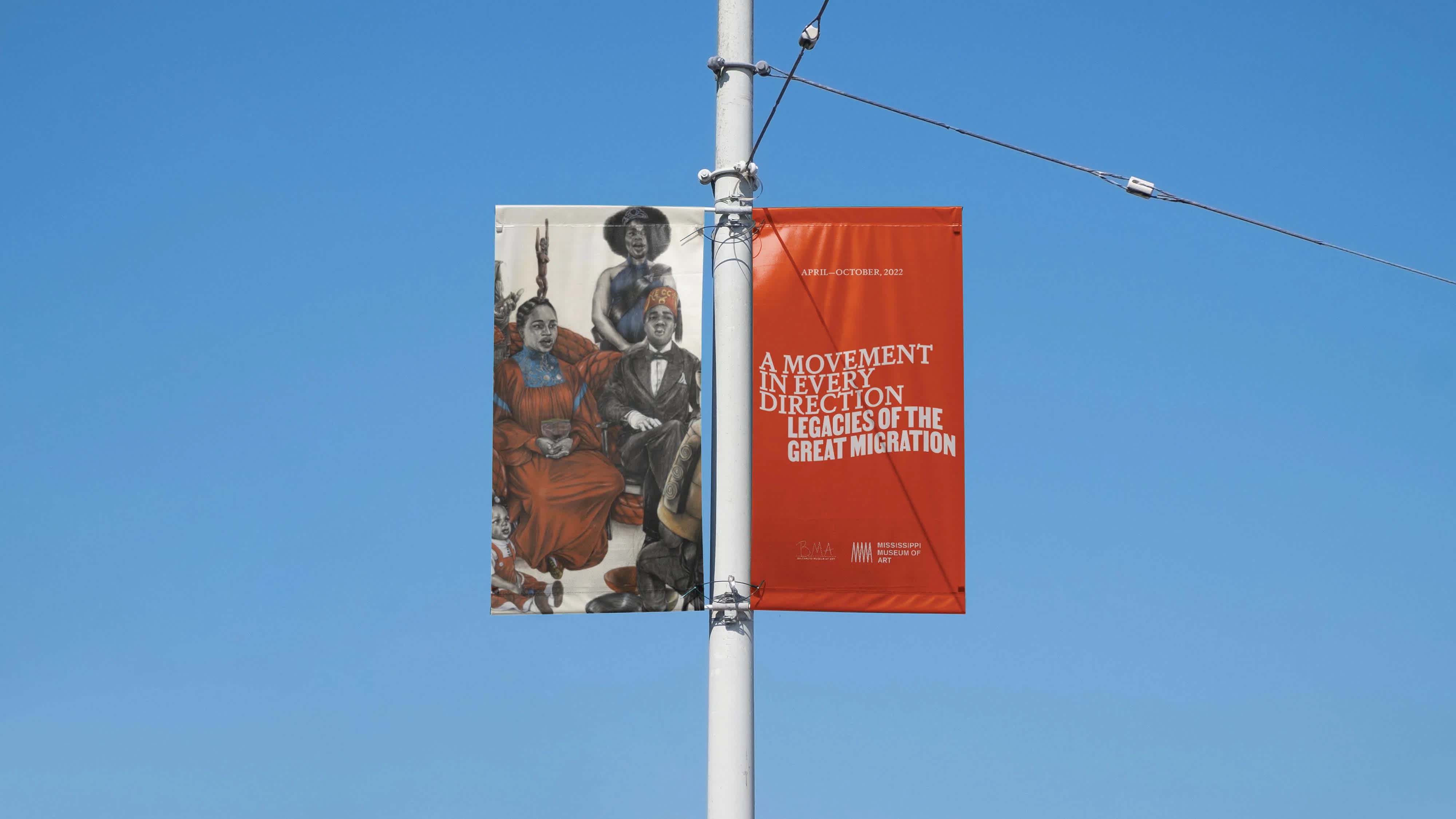 A lamp post with two banners on either side. The left banner features artwork. The right banner is an advertisement for an exhibition. The sky behind is bright blue and clear.